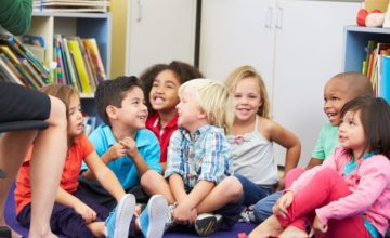 A group of preschoolers sit on the carpet together in their classroom.