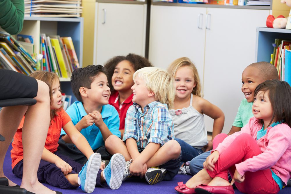 A group of preschoolers sit on the carpet together in their classroom.