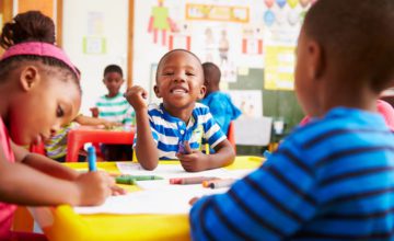 Children in preschool, three infants coloring, one smiling directly at camera