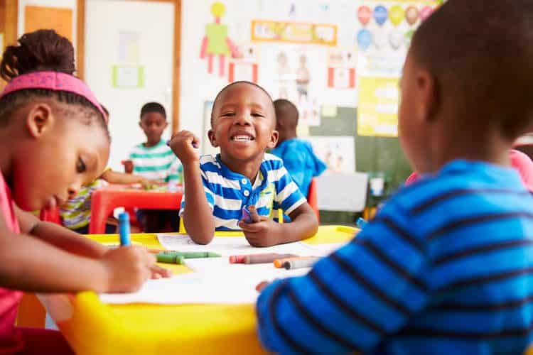 Children in preschool, three infants coloring, one smiling directly at camera