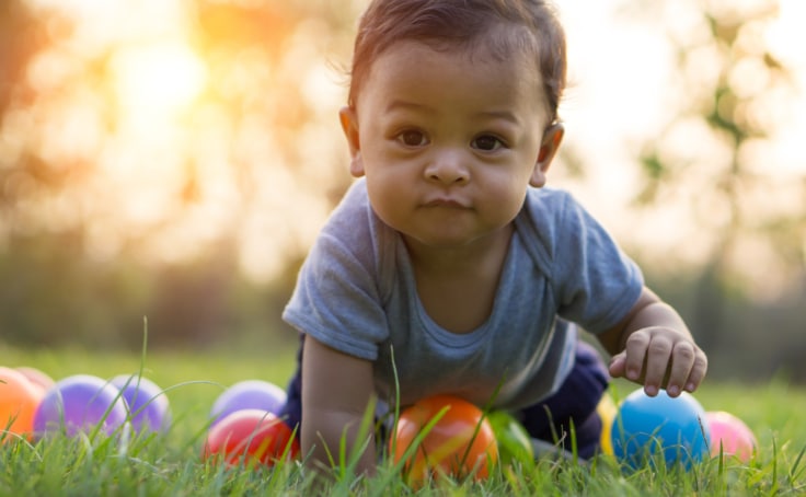 An infant outside in the grass crawling with colored toys around