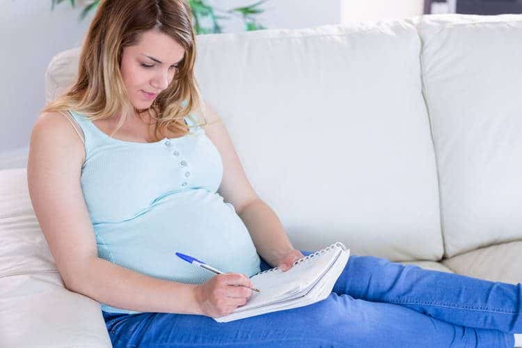 A pregnant woman sits on a white couch writing in a journal.