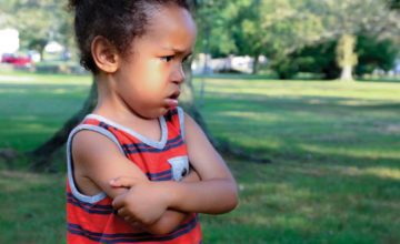 A small child stands with arms crossed across chest and looks upset.