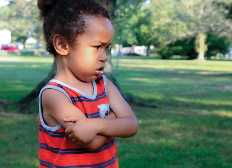 A small child stands with arms crossed across chest and looks upset.