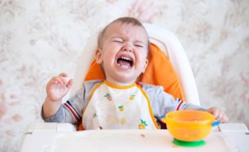 Baby sitting in high chair eating from a bowl and crying