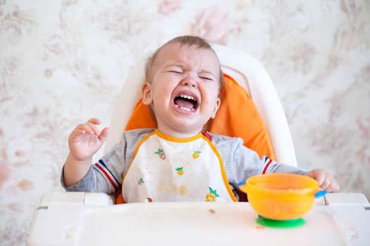 Baby sitting in high chair eating from a bowl and crying