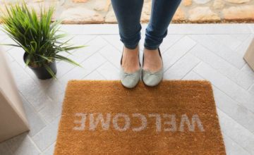 Feet on edge of welcome mat on a doorstep with cardboard boxes and a potted plant