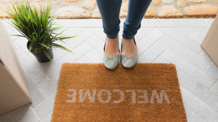 Feet on edge of welcome mat on a doorstep with cardboard boxes and a potted plant