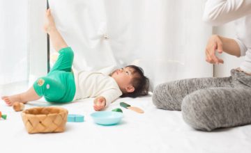 Toddler laying o her back and putting her leg up against window curtain