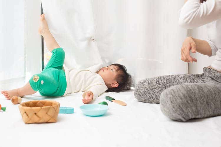 Toddler laying o her back and putting her leg up against window curtain