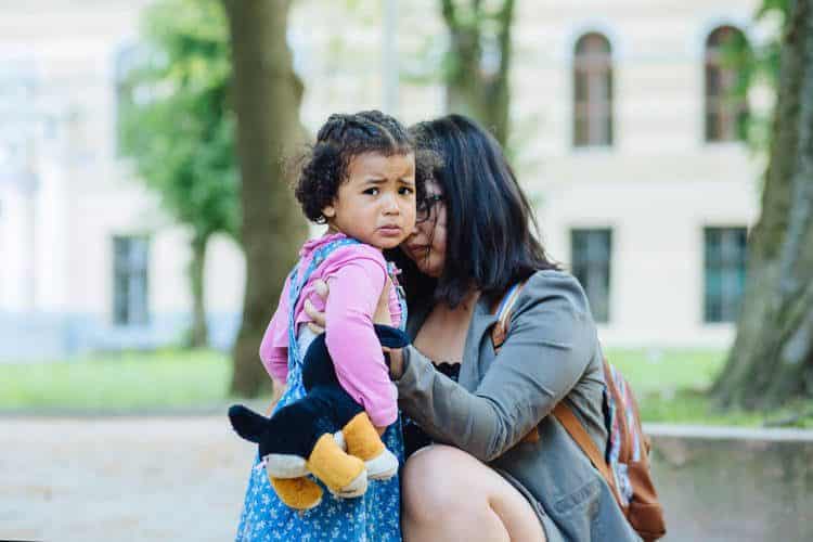 Mom holding fussy toddler who is crying, standing outdoors.