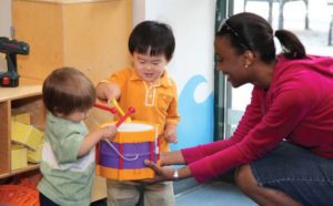 A child care educator holds a toy drum for two children to play with.