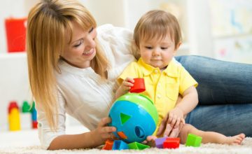 A mother sits on the ground with her child and plays with a shape toy.