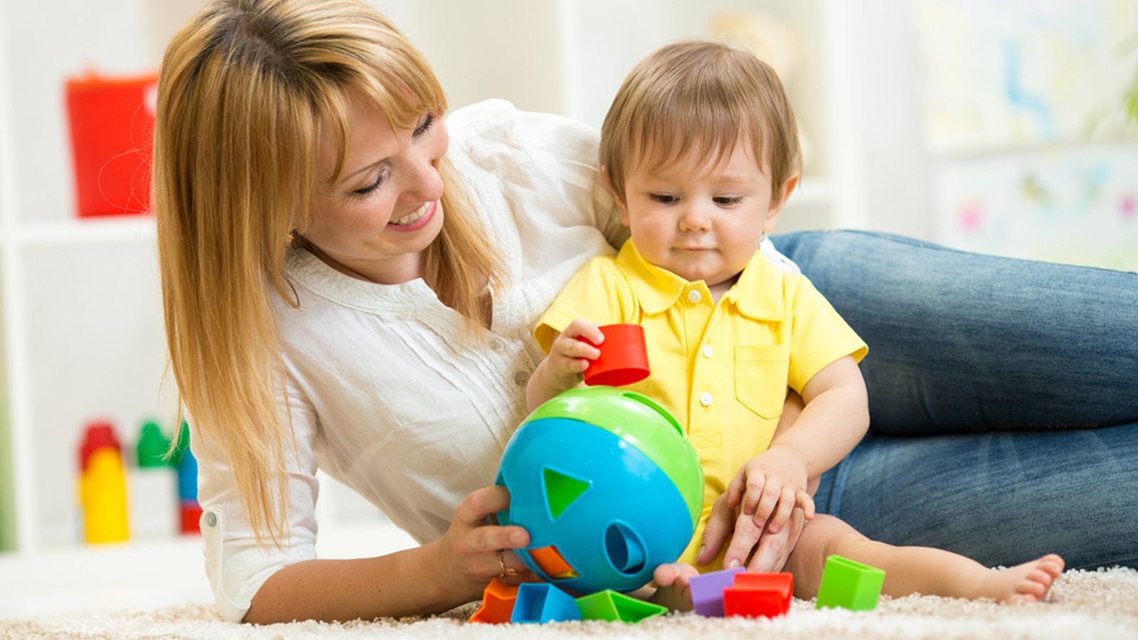 A mother sits on the ground with her child and plays with a shape toy.