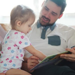 A toddler points to the book her father is reading to her.