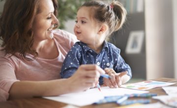 A mother and daughter look at each other while drawing with crayons.