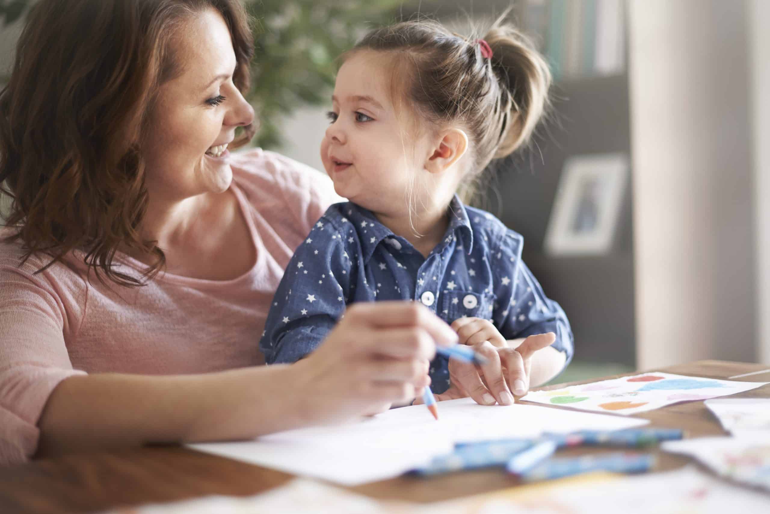 A mother and daughter look at each other while drawing with crayons.