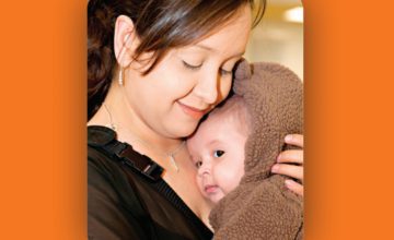 mother smiling holding baby in brown coat close to chest