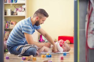 father sitting cross-legged on floor changing baby diaper with toys around