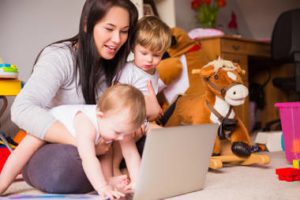 woman holding two children on floor looking at laptop