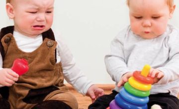 Two infants sitting with toys in hand, one crying one focused on the toy