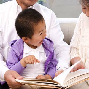 Child pointing at book while sitting on man's lap and looking at another adult