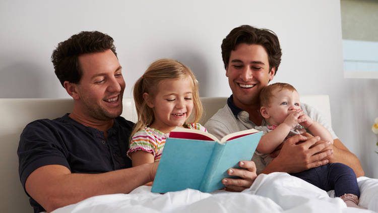 Two smiling men read a book to a child and infant