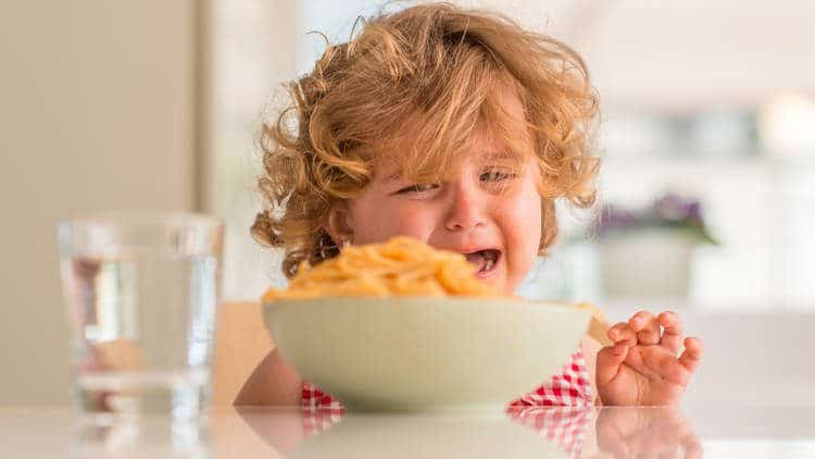 Crying toddler sitting at table with bowl of food and glass of water.