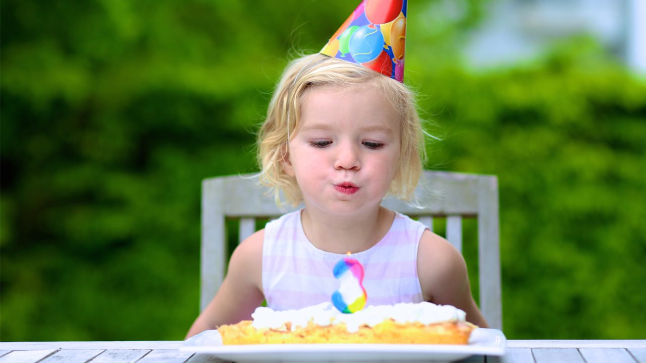 little girl sits at table and blows out birthday candle