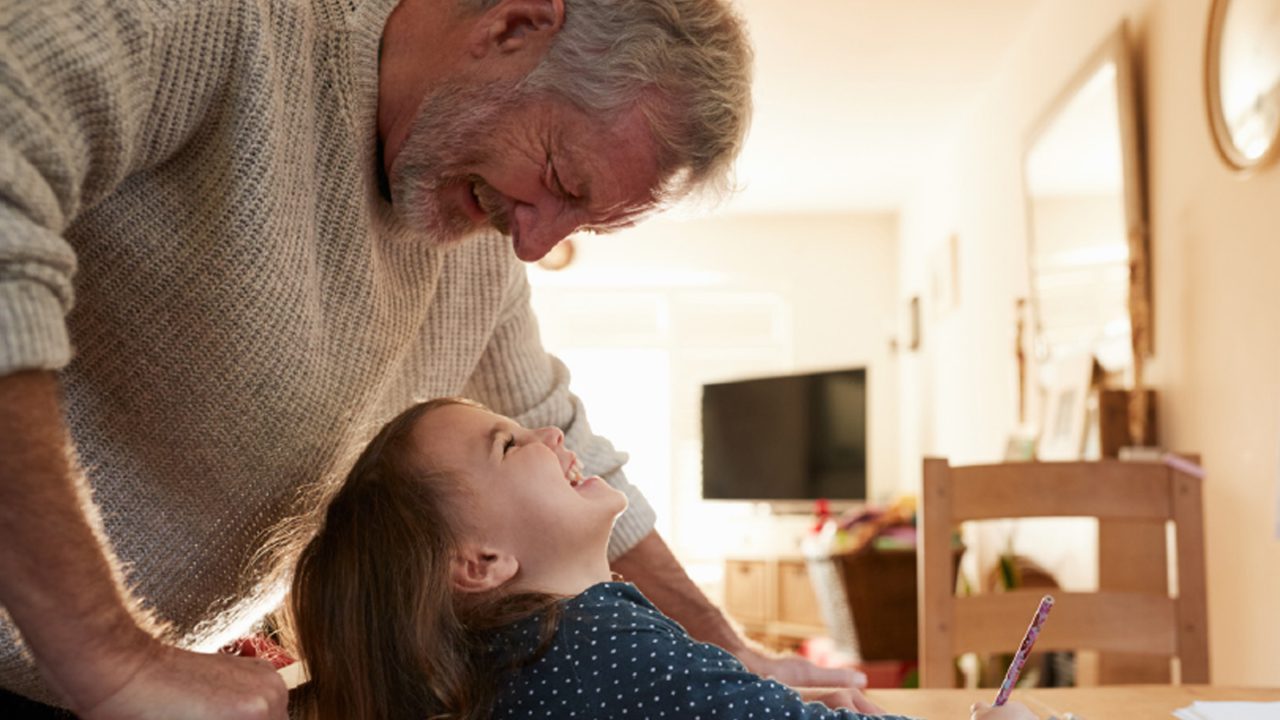 A child sitting and drawing at a table looks up at her grandfather, both smiling.