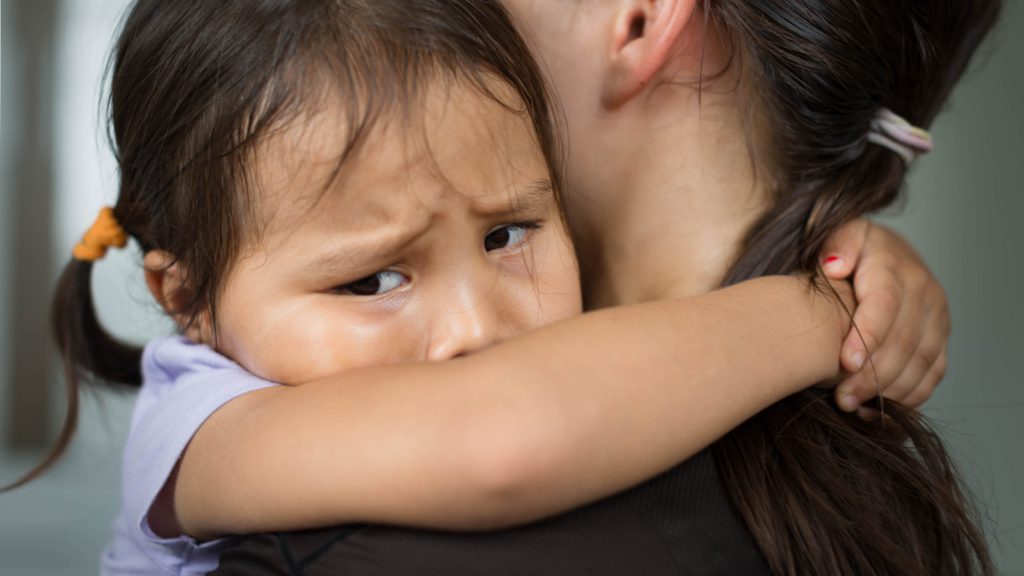 A crying child hugs her mom around the neck.