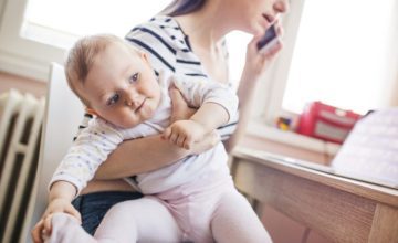 A mother holds her baby under her arm while she talks on a cell phone and looks at a laptop.