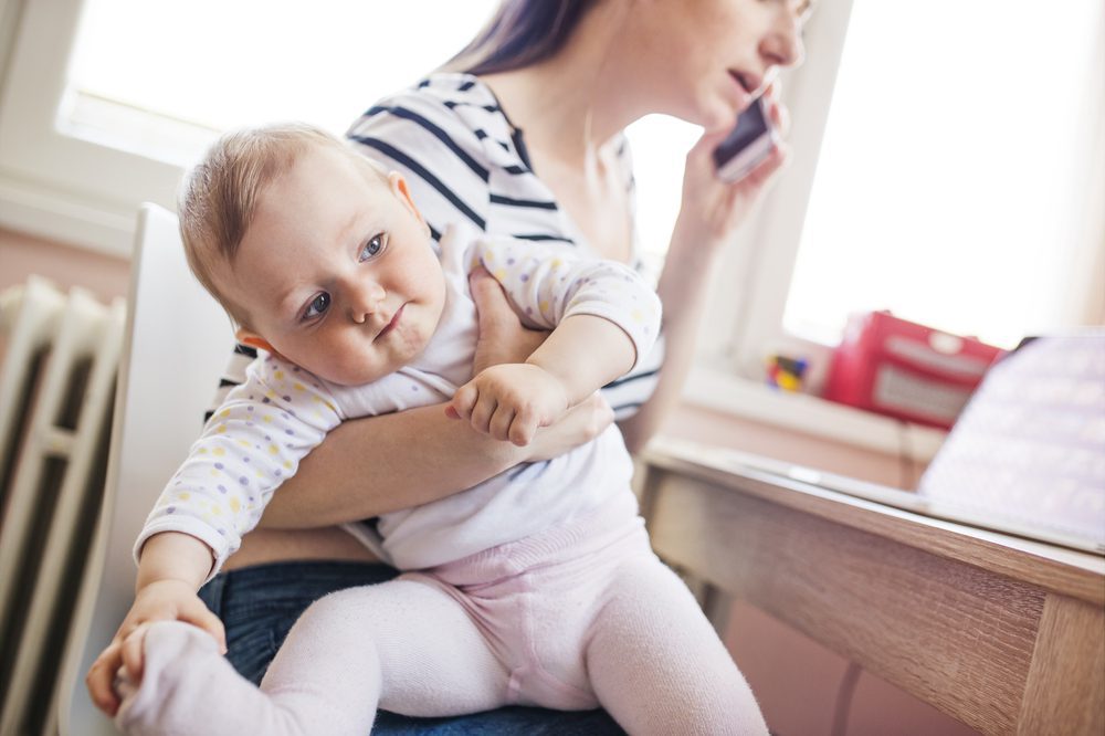 A mother holds her baby under her arm while she talks on a cell phone and looks at a laptop.