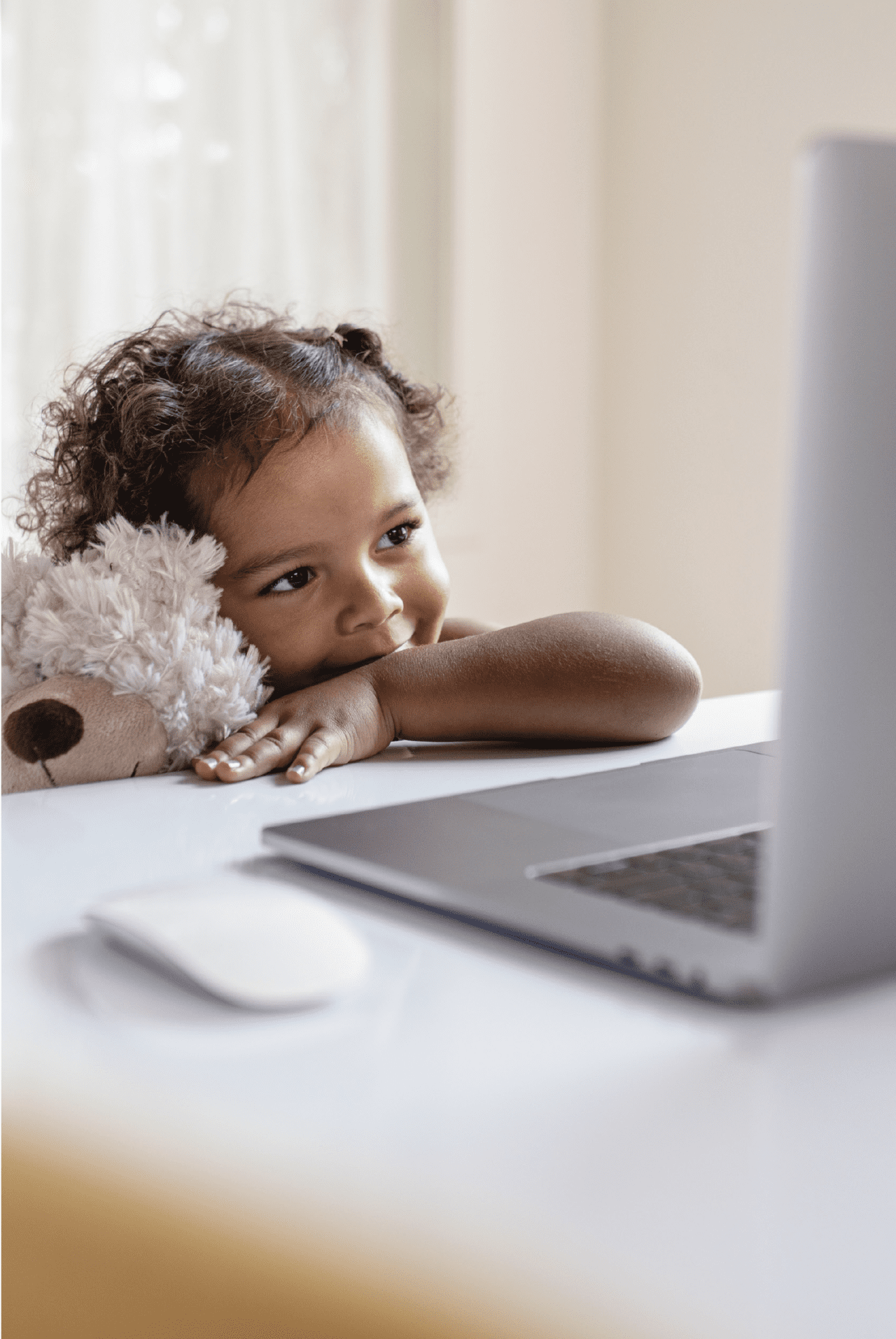 Small girl holds a teddy bear and looks at a laptop screen.