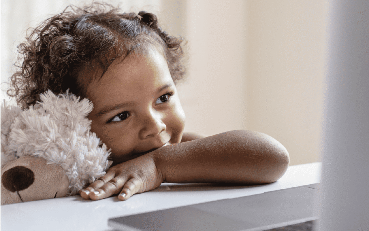 Small girl holds a teddy bear and looks at a laptop screen.