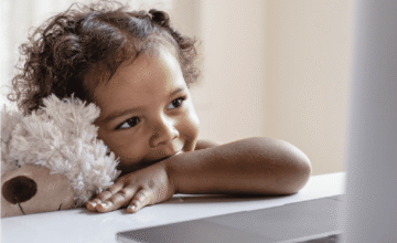 Small girl holds a teddy bear and looks at a laptop screen.
