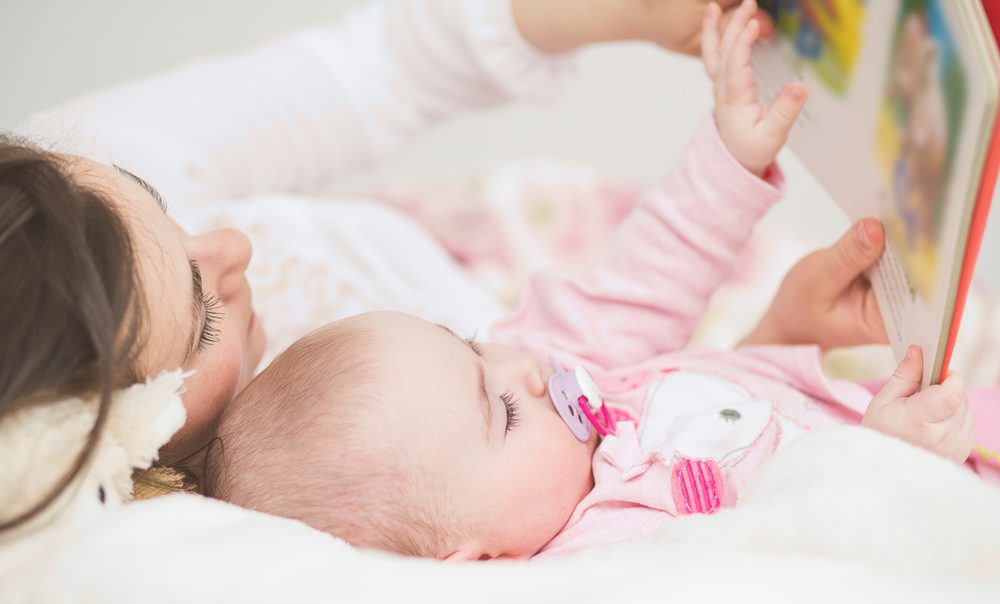 A mother and baby lie in bed looking at a storybook.