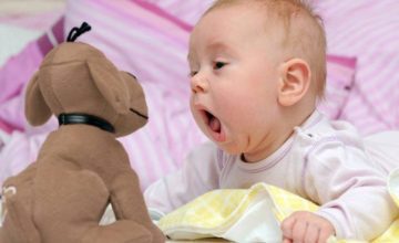 A baby with its mouth open staring at a stuffed toy dog in front of pillows