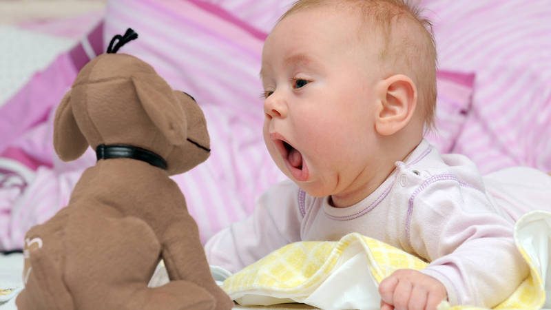 A baby with its mouth open staring at a stuffed toy dog in front of pillows