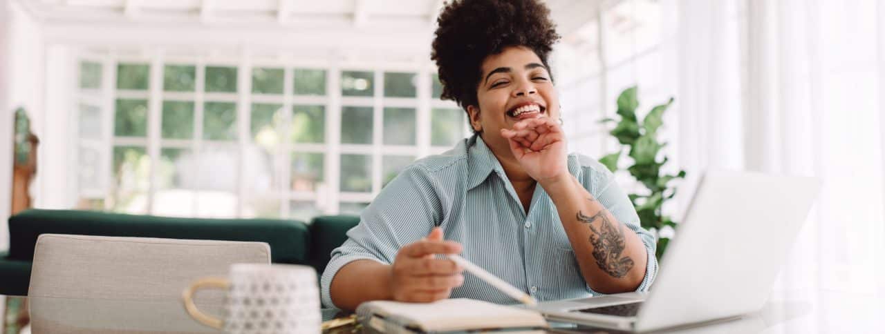 Wide angle shot of happy female sitting at desk looking at camera. Attractive woman smiling while working from home.