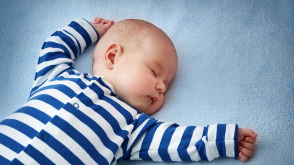 A baby wearing blue and white striped pajamas sleeping with arms above head. 