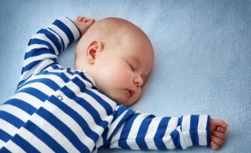 A baby wearing blue and white striped pajamas sleeping with arms above head.