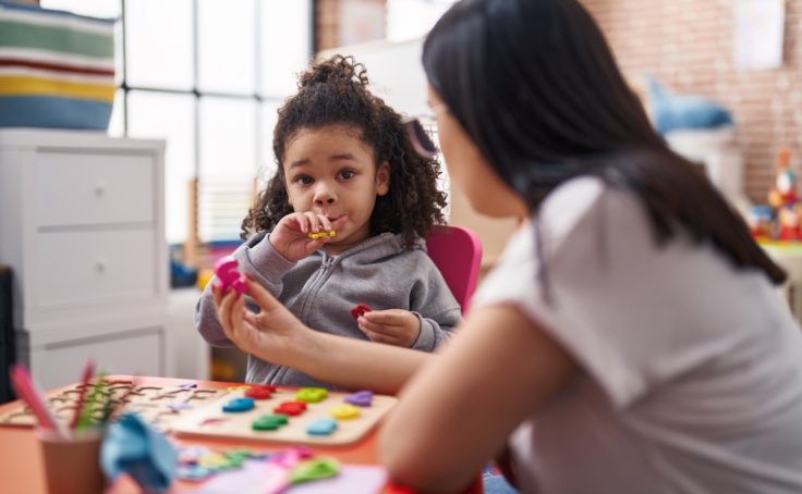 Teacher and toddler playing with maths puzzle game sitting on table at kindergarten