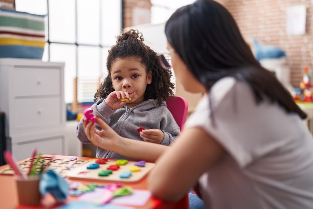 Teacher and toddler playing with maths puzzle game sitting on table at kindergarten