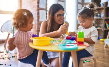 Young beautiful teacher and toddlers playing meals using plastic food and cutlery toy at kindergarten