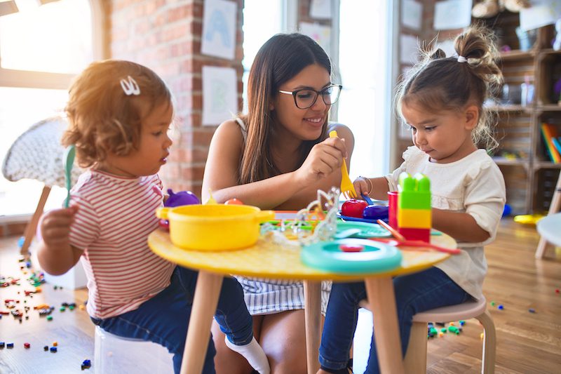 Young beautiful teacher and toddlers playing meals using plastic food and cutlery toy at kindergarten