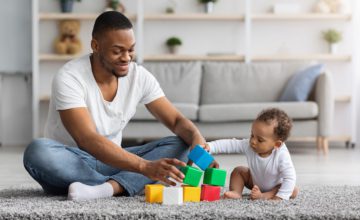 Happy Young Black Father Playing With His Little Infant Baby Son At Home, African American Dad And His Cute Toddler Male Child Stacking Building Blocks While Sitting Together On Floor In Living Room