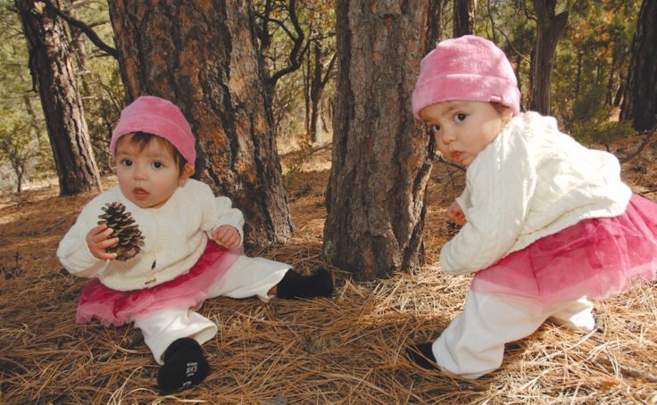Twin girls wearing matching pink and white outfits sit together near trees.