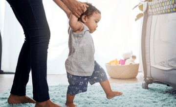 A baby boy learning to walk. Arms are held up by a parent for stability.