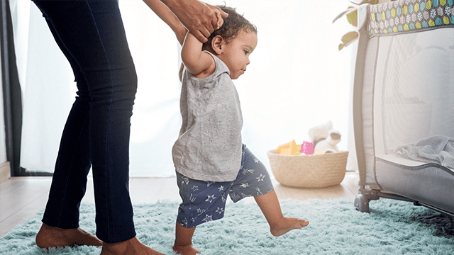 A baby boy learning to walk. Arms are held up by a parent for stability.